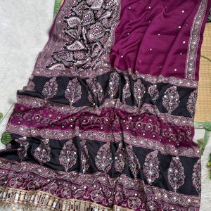 Op de afbeelding: Een dieppaarse sari met een zwarte rand met ingewikkelde witte en paarse paisley-patronen. De stof is versierd met kleine reflecterende spiegelaccenten over het hele lichaam en een decoratieve franje met hangende spiegelstukjes.