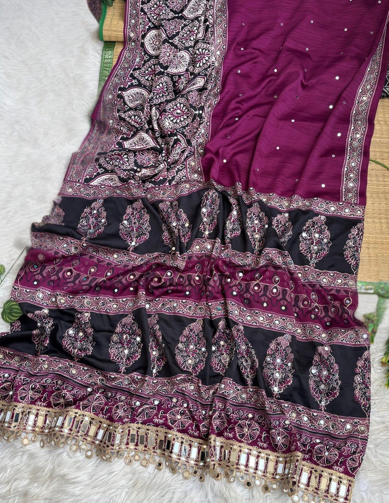 Op de afbeelding: Een dieppaarse sari met een zwarte rand met ingewikkelde witte en paarse paisley-patronen. De stof is versierd met kleine reflecterende spiegelaccenten over het hele lichaam en een decoratieve franje met hangende spiegelstukjes.