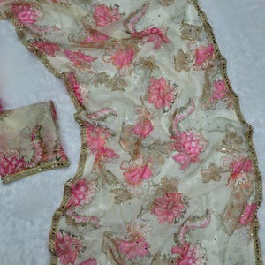 May include: A cream-coloured, sheer fabric featuring a floral pattern in pink and brown. The fabric is embellished with sequins and has a gold-coloured border. A matching piece of fabric is also present.