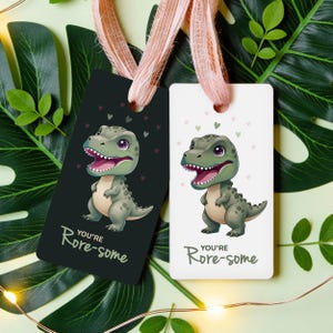 Könnte beinhalten: Zwei Geschenkanhänger mit einer Cartoon-Dinosaurier-Illustration. Einer ist schwarz, der andere weiß. Beide haben ein rosa Band und den Text "You're Rore-some". Der Dinosaurier ist grün mit einem rosa Mund und lila Augen.