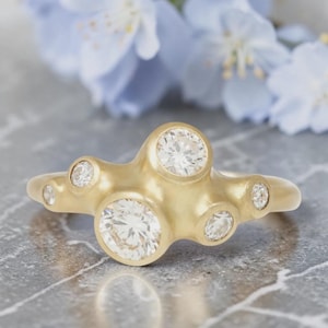 Op de afbeelding: Een gouden ring met zeven heldere, ronde diamanten. De ring heeft een uniek, organisch ontwerp met een vloeiende, onregelmatige vorm. De ring is geplaatst op een grijze marmeren ondergrond met zachte blauwe bloemen op de achtergrond.