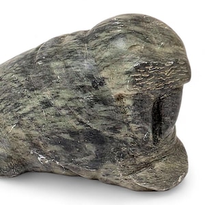 May include: A carved stone sculpture in shades of gray and green. The sculpture has a rounded shape with a textured surface, possibly depicting a stylized figure or animal. The stone has a natural, marbled appearance.