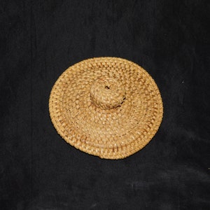 Puede incluir: Una tapa redonda de paja tejida con un centro elevado. La tapa es de color marrón claro y tiene un aspecto natural y rústico.