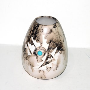 May include: A rounded ceramic vase with a cream-colored base featuring black, web-like smoke patterns. The surface is decorated with white geometric shapes and a single, centered, circular turquoise stone inlay on the front side.