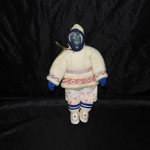 May include: A handmade doll dressed in traditional Inuit clothing. The doll wears a white parka with blue and red accents, blue mittens, and patterned pants. The doll's hood is trimmed in blue, and the face is a dark color.