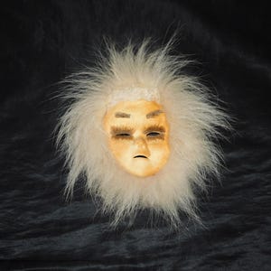 May include: A white fur mask with a painted face. The face is light brown with closed eyes and a neutral expression.