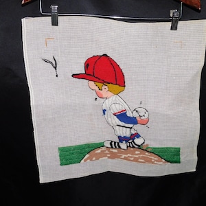 May include: A needlepoint canvas depicting a cartoon baseball player wearing a red hat, blue and white jersey, and white trousers. The player is holding a baseball and standing on a brown dirt mound.