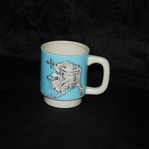 May include: A ceramic mug with a light blue exterior and a white interior and base. The mug features a detailed gray and white map of Alaska. The handle and base are white.