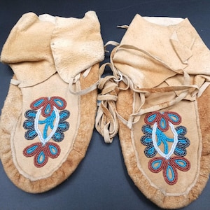 May include: A pair of tan leather moccasins with a decorative beaded design in blue, red, and white. The moccasins have a folded-over cuff and are tied with leather laces. The design is symmetrical and floral.