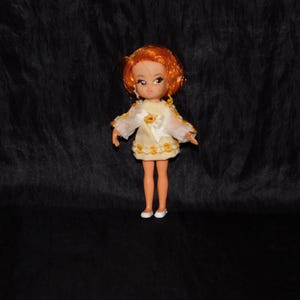 Puede incluir: Una muñeca vintage con pelo naranja que lleva un vestido blanco con flores amarillas y un lazo blanco. La muñeca lleva zapatos blancos.