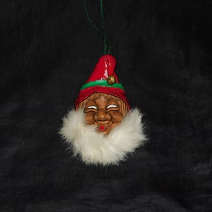 May include: A handmade Christmas ornament featuring a whimsical, brown-skinned elf face with a white fur beard and a red hat with green trim. The ornament has a bell attached to the hat.