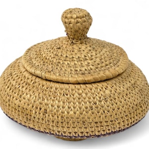 Puede incluir: Una cesta redonda tejida con tapa. La cesta está hecha de fibras de color natural y tiene un botón en la parte superior de la tapa. La cesta tiene aproximadamente 15 cm de diámetro y 10 cm de altura.