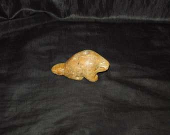 May include: A hand-carved stone sculpture of a beaver, with a brown and tan color scheme. The sculpture is sitting on a black surface.