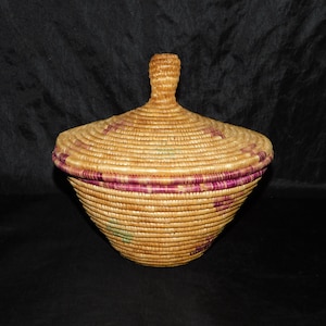 May include: A woven basket with a lid, featuring a natural straw color with purple accents. The basket has a round, slightly conical shape.