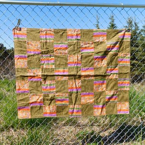 May include: A patchwork quilt with a repeating pattern of olive green squares and rectangular stripes in orange, purple, and green. The quilt is hanging on a chain-link fence outdoors on a sunny day.