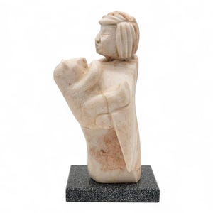 May include: A carved stone sculpture of a person holding an object, possibly a child. The sculpture is light beige with reddish-brown accents and sits on a dark gray, speckled base. The figure has a serene expression.