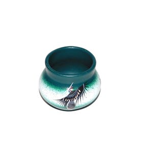 May include: A small, round, teal-colored ceramic pot with a wide opening. The pot features a hand-painted design of a bird with black and white feathers. The base of the pot has a speckled white and green pattern.