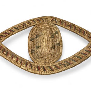 Puede incluir: Cesta tejida en forma de ojo. El óvalo central está rodeado por un marco más grande en forma de ojo. La cesta está hecha de fibras de color natural con acentos rojos y verdes. El diseño es simétrico y parece hecho a mano.