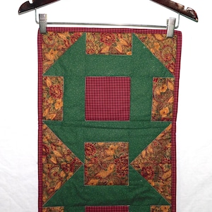 May include: A quilted table runner with a green, red, and brown patchwork design. The runner features a repeating pattern of squares and triangles.