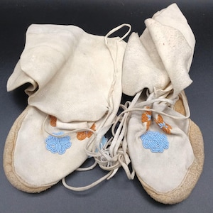 Może przedstawiać: Para kremowych skórzanych mokasynów z niebieskimi i pomarańczowymi wzorami kwiatowymi z koralików. Moccasins mają miękką, zamszową teksturę i są wiązane skórzanymi sznurowadłami. Wierzchy są złożone.