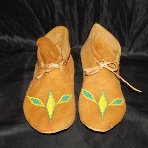 May include: A pair of tan leather moccasins. Each moccasin features a beaded design in yellow and teal. The moccasins have a soft sole and a leather tie at the ankle. The moccasins are approximately 6 inches long.