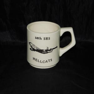 May include: Cream-colored ceramic mug with a black graphic of an airplane and the text "38th SRS HELLCATS." The mug has a square handle and a simple, classic design, suitable for coffee or tea.