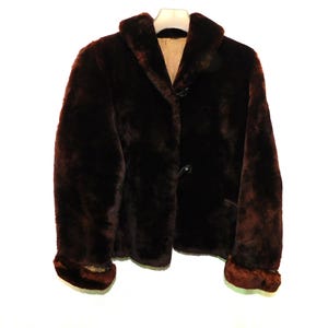May include: A dark brown fur jacket with a classic collar and button closure. The jacket has a plush texture and is hanging against a white background. The cuffs and collar have a slightly lighter brown trim.