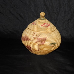 May include: A woven basket with a lid, featuring a brown, red, and green geometric design. The basket is made of natural materials and has a round shape.