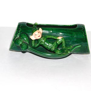 May include: A green ceramic planter shaped like a log with a green elf figurine lying on top. The elf has a pointed hat and is holding a leaf.