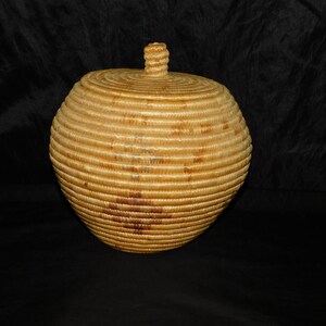 May include: A round, lidded basket with a light brown hue. The basket is made of woven material, with horizontal bands creating a textured surface. The lid has a small, spiraled knob. The basket is set against a dark background.