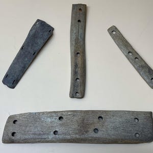 May include: Four weathered, gray wooden planks with multiple drilled holes. The planks vary in size and shape, with one long, rectangular piece and three smaller, narrower pieces. The wood has a textured appearance, suggesting age or exposure to the elements.