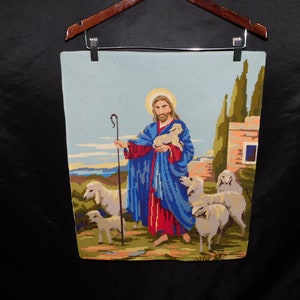 May include: A needlepoint wall hanging depicting Jesus as the Good Shepherd. He is holding a lamb in his arms and has a staff in his left hand. He is surrounded by sheep in a green field.
