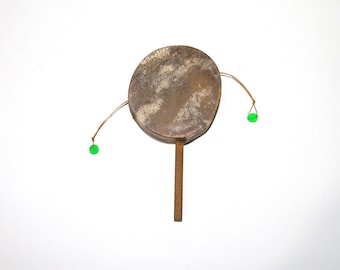 May include: A small, round percussion instrument with a brown, textured drumhead. It has a wooden handle and two strings with green beads attached. The drumhead has a mottled appearance, and the instrument is set against a white background.