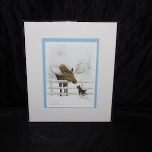 May include: A framed print featuring a moose and a small black dog in a snowy landscape. The moose stands behind a wooden fence, facing the dog. The artwork has a blue border and is set against a white background.