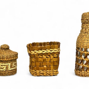 Op de afbeelding: Drie handgemaakte geweven items: een kleine pot met deksel en geometrisch patroon, een vierkante mand en een flesvormig item. Alle zijn gemaakt van natuurlijk gekleurd geweven materiaal, met een rustieke uitstraling. De fles heeft een roosterontwerp.
