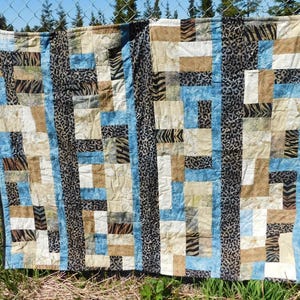 May include: A handmade quilt with a patchwork design. The quilt features a mix of blue, tan, and cream-colored rectangular blocks, accented with zebra and leopard print fabric. The quilt is hanging outdoors, with a chain-link fence and trees in the background.