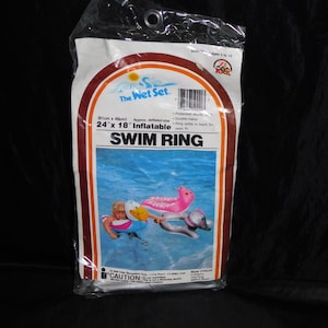 Puede incluir: Un paquete de plástico transparente que contiene un anillo de natación inflable. El empaque presenta el texto The Wet Set, SWIM RING y 61cm x 46cm Inflatable. Muestra imágenes de niños usando anillos de natación con formas de animales.