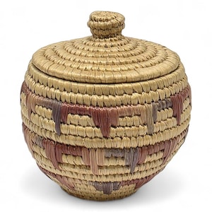 以下が含まれることがあります： 天然繊維で織られた、ふた付きの丸いバスケット。バスケットは、茶色と赤の色合いの幾何学模様を繰り返しています。ふたにはノブハンドルが付いています。バスケットは、おそらく保管または装飾に使用されます。