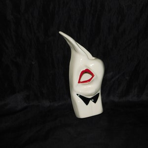 May include: White ceramic vase shaped like a face with red lips and a black bow tie.