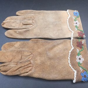 Op de afbeelding: Een paar bruinleren handschoenen met geborduurde bloemenmanchetten. De manchetten hebben witte geschulpte randen en kleurrijk kralenwerk in blauw, rood, groen en wit, wat een decoratief accent creëert.