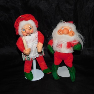 Puede incluir: Un par de adornos navideños vintage que representan a Papá Noel y a la señora Claus. El adorno de Papá Noel lleva un traje rojo con ribetes blancos y una barba blanca. El adorno de la señora Claus lleva un vestido rojo con un delantal blanco y un gorro blanco. Ambos adornos tienen botas de fieltro verde.