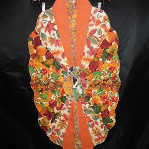 May include: A fall-themed table runner with a scalloped edge. The runner is made of fabric with a patchwork design featuring orange, brown, and white leaves, and a central orange stripe.