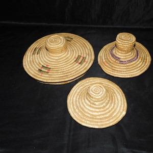 May include: Three woven straw hats of varying sizes. The hats have a natural beige color with decorative bands of green, red, and purple. The hats are arranged on a dark surface.