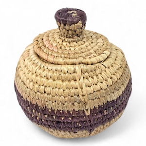 Może przedstawiać: Okrągły kosz z pokrywką, tkany z naturalnych i fioletowych włókien. Kosz ma okrągły wzór z gałką na pokrywce. Podstawa kosza ma fioletowy pas.