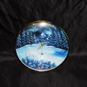 Pode incluir: Um relógio redondo de vidro com moldura dourada. O mostrador do relógio apresenta uma cena de inverno pintada com uma paisagem nevada, árvores e montanhas. A assinatura do artista, "Bob Ross", é visível no canto inferior direito.