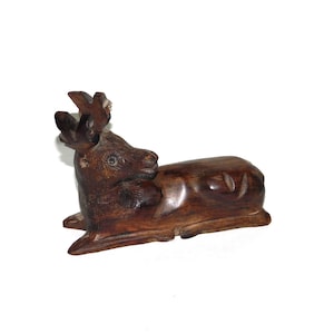 May include: A dark brown wooden carving of a deer lying down with its head turned to the side. The deer has antlers and detailed facial features.