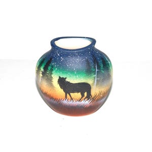 May include: A round ceramic vase with a painted landscape scene. The design features a black wolf silhouette against a sunset, with trees and a starry night sky. The vase has a dark blue rim and a white interior.