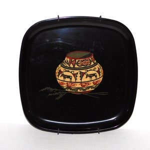 Puede incluir: Bandeja cuadrada negra con esquinas redondeadas. La bandeja presenta una ilustración colorida y detallada de una olla nativa americana con diseños geométricos y figuras de animales. La bandeja tiene asas de metal.