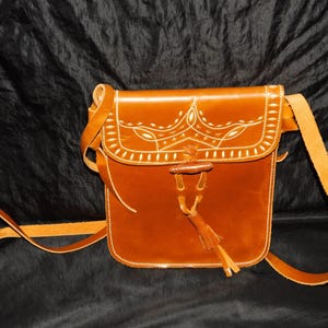 May include: A brown leather crossbody bag with white stitching and a tassel. The bag has a flap closure and a long adjustable strap.