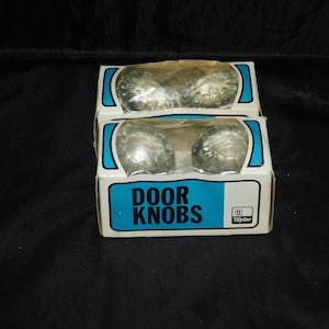 May include: Two boxes of door knobs are stacked on top of each other. Each box has a blue and white design and the words "DOOR KNOBS" printed on the front. The door knobs are silver and are visible through a clear plastic window.